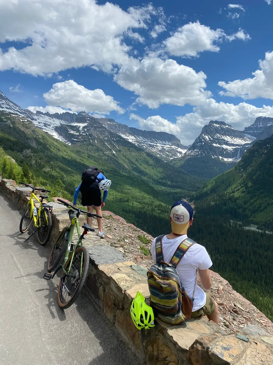 Cyclists resting at a stone wall overlook on the Going-to-the-Sun Road with peaks behind