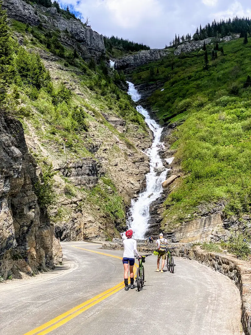 Cyclists riding past the Weeping Wall waterfall pouring onto the Going-to-the-Sun Road
